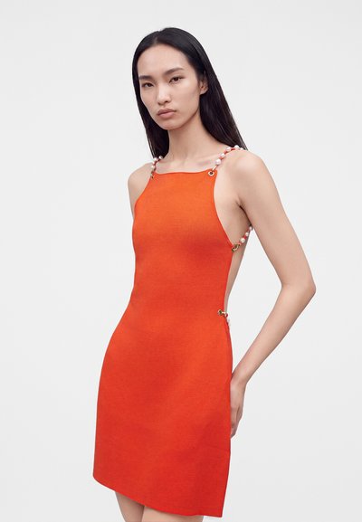 Femme portant une robe orange sans manches avec des côtés ouverts à bretelles perlées, debout devant un fond gris clair uni.