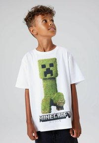 Valkoinen t-paita, jossa on vihreä pikselöity Creeper-hahmo Minecraftista. Paidan alaosassa on musta "Minecraft-elokuva" -logo.