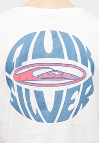 Valge särgi tagakülg, millel on suur sinine ümmargune kiri "Quiksilver", mis ümbritseb keskel punast ja sinist stiliseeritud lainelogo.