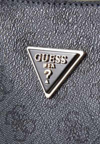 Superficie nera testurizzata con un logo triangolare metallico che presenta la scritta "GUESS USA" e un dettaglio a punto interrogativo.