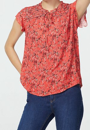 Kvindelig iført en rød blomstret bluse med korte ærmer og snørredetalje, parret med blå denim jeans, stående med den ene arm hævet.