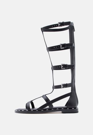 Sandale gladiateur noire montante jusqu'au genou avec plusieurs brides réglables à boucle et des clous argentés le long du bord de la semelle.