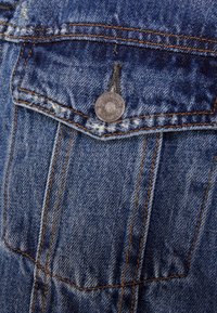 Modrá denimová tkanina s kapsou, která ukazuje detaily ve formě stříbrného knoflíku. Má viditelné švy a vybledlou texturu.