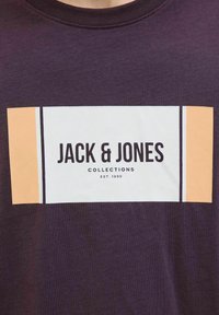 Camiseta de algodón morada con un gráfico rectangular en blanco y melocotón que presenta "JACK & JONES" y "COLLECTIONS EST. 1990" en texto negro en negrita.