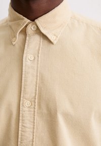 Chemise en velours côtelé beige avec un col boutonné, présentant une texture rayée horizontale et de petits boutons ronds le long de la patte de boutonnage.