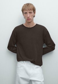 Bruine blouse met lange mouwen en een ronde halslijn, gecombineerd met witte broek. De blouse heeft een ontspannen pasvorm en een zachte textuur.