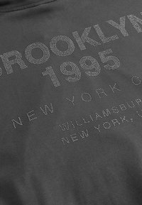 Harmaa puuvillapaita, jossa on kohotettuja hopeisia strasseja, jotka muodostavat sanat "BROOKLYN 1995" ja "NEW YORK CITY" eri kokoisina ja fontteina.