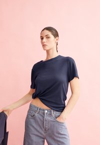 T-shirt bleu marine à manches courtes avec une coupe décontractée, légèrement froncée à la taille, associé à un jean en denim délavé. Fond rose.