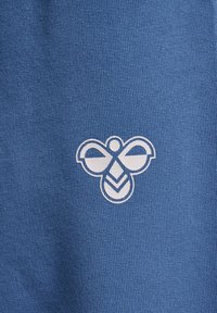 Blå bomullstyg med en vit bi-logotyp med stiliserade vingar och ett geometriskt mönster. Texturen är slät och stickad.