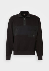 Pull noir à manches longues avec fermeture éclair au quart, bande de tissu horizontale et poche poitrine avec rabat, suspendu sur fond blanc.