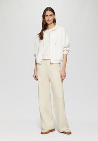 Sweat à fermeture éclair blanc, sweatshirt ajusté crème, et pantalon large beige clair. Tissu lisse, design minimaliste, avec un motif de cœur sur le pantalon.