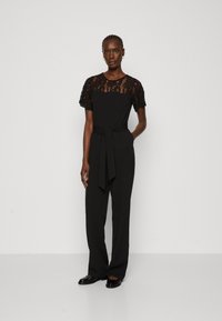 Combinaison noire avec un haut en dentelle à manches courtes, une taille ceinture et un pantalon droit. Tissu lisse avec un léger éclat.