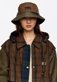 Chaqueta de cuadros marrón con una mezcla de acentos verdes, azules y rojos; cuenta con un cuello forrado de piel y un parche con el logo. Combinada con un sombrero tipo cubo marrón y verde.