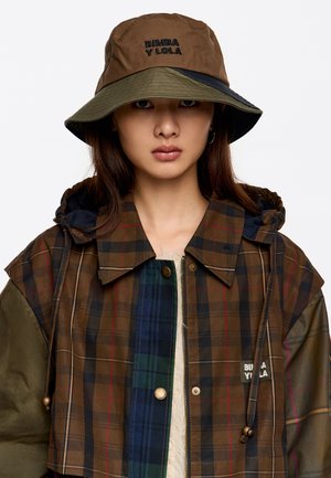 Chaqueta de cuadros marrón con una mezcla de acentos verdes, azules y rojos; cuenta con un cuello forrado de piel y un parche con el logo. Combinada con un sombrero tipo cubo marrón y verde.