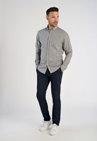 Camicia grigia con bottoni e tasca, pantaloni blu scuro e sneakers bianche. La camicia presenta un tessuto texturizzato e un colletto classico.