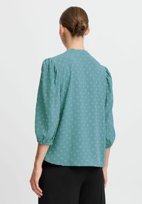 Blusa teal in tessuto leggero con maniche a sbuffo, scollo rotondo e piccoli motivi floreali su tutto il capo. Abbinata a pantaloni neri.