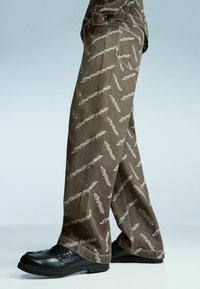 Pantaloni largi maro cu un design repetat de model gri deschis. Au buzunare laterale și sunt purtați cu pantofi negri.