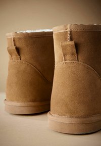 Smeđe suede čizme do gležnja s mekanym bijelim podstavljanjem, okruglog oblika, sa šivenim detaljima i kolektorskim trakama straga za lakše obuvanje.