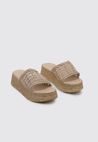 Sandalias de slide de color beige tejidas con una tira ancha, plataforma de yute natural y superficies texturizadas. Diseño resistente para comodidad y estilo.
