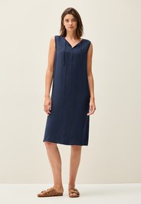 Marineblauwe mouwloze jurk met een V-hals en trekkoord. De stof is lichtgewicht met een gestructureerde afwerking. Het model draagt bruine sandalen met een polka dot-print.