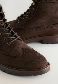Bruine suède laarzen met ingewikkelde brogue-details, ronde punt en stevige veters, tegen een neutrale achtergrond.