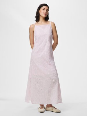 Femme portant une robe longue sans manches rose pâle avec une broderie délicate, debout face à l'avant, fond neutre.