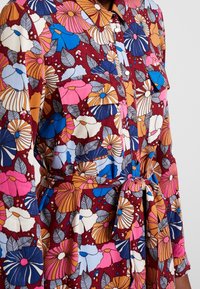 Blommig klänning med en mörkröd bakgrund och flerfärgade mönstrade blommor. Den har en krage, knappstängning fram och en knytdetalj i midjan.