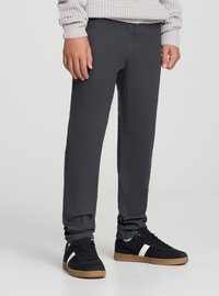 Persona con pantaloni scuri aderenti, sneakers nere con striscia bianca e maglione lavorato a maglia di colore grigio chiaro, in piedi su uno sfondo semplice.