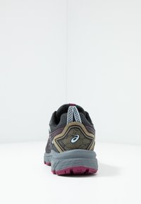 Chaussure de course noire et grise avec une tige texturée, des accents couleur beige et une semelle en caoutchouc violette. Les lacets sont blancs avec une languette arrière de traction.