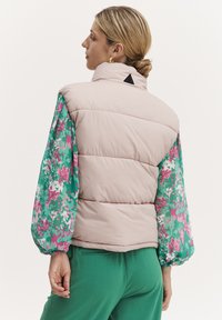 Vestă umplută de culoare roz deschis cu guler înalt, având mâneci voluminoase cu model floral verde și un detaliu cu logo negru pe spate.