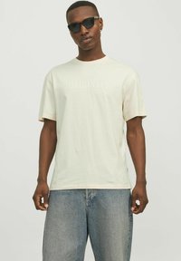Un tricou scurt de culoare bej deschis, cu inscrip�ția "ORIGINALS" embosată în centru, purtat cu blugi largi de denim albastru.
