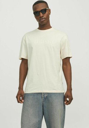 Camiseta de manga corta en color beige claro con la palabra "ORIGINALS" estampada en el centro, combinada con jeans de mezclilla azul holgados.