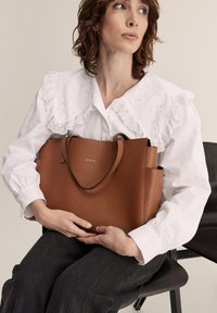 Sac fourre-tout en cuir marron avec une finition texturée, deux poignées et un branding subtil. Le modèle porte un chemisier blanc avec un col en dentelle, assis sur une chaise.