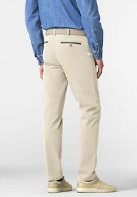 Man in beige broek met een bruine riem, een blauwe blouse met lange mouwen en beige sneakers, staande tegen een effen lichte achtergrond.