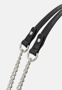 Černé kožené popruhy kabelky připevněné k stříbrným řetězovým článkům s kovovými nýty na bílém pozadí.