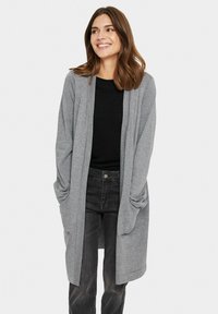 Cardigan grigio a maglia lunga con apertura frontale, maniche larghe e tasche. Abbinato a una maglietta nera e jeans scuri, che mette in evidenza una vestibilità comoda.