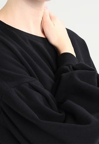 Sudadera negra de cuello redondo con mangas voluminosas recogidas y puños de canalé. Textura de tela suave, mostrada en una persona con una mano en el cuello.