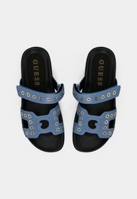 Οι μπλε σανδάλες slides διαθέτουν δύο λουράκια με χρυσά μεταλλικά διακοσμητικά, μια κεντρική διακοσμητική αγκράφα και μαύρη σόλα με το λογότυπο "GUESS".