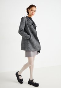 Manteau en mélange de laine gris avec des boutons croisés, arborant un large col. Le modèle porte une jupe grise et des chaussures Mary Jane à plateforme noires.