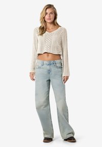 Krämfärgad stickad korttröja med V-ringning och diamantmönster, kombinerad med ljusblå vida jeans och mörka sandaler.