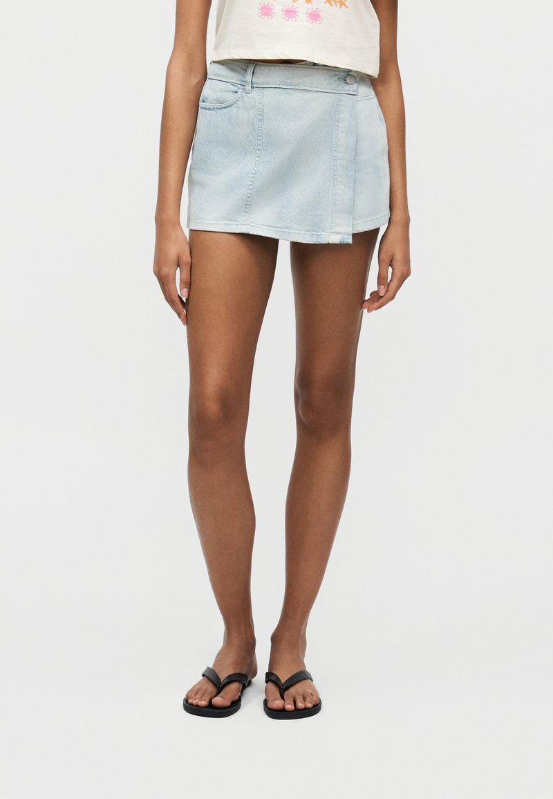 Γυναίκα που φοράει μια κοντή φούστα από ανοιχτό μπλε τζιν, μπεζ κοντή μπλούζα με πορτοκαλί και ροζ σχέδια, και μαύρα σαγιονάρες σε απλό φόντο.