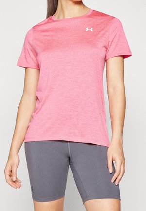 Frau trägt ein pinkfarbenes kurzärmeliges Under Armour T-Shirt und graue eng anliegende Shorts und steht vor einem einfarbigen Hintergrund.