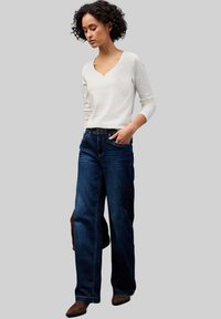 Top lungo a maniche lunghe bianco con righe abbinato a jeans di denim blu scuro dall'fit morbido. Il modello sta in piedi con una mano in tasca, indossando stivaletti marroni alla caviglia.