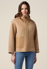 Giacca in misto lana color sabbia con colletto alto, zip completa e due tasche frontali. Presenta una forma squadrata e maniche larghe per un fit rilassato.