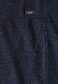 Mörkblått tyg med en strukturerad yta, med en liten metalletikett märkt "CECIL" sydd ovanför en söm.
