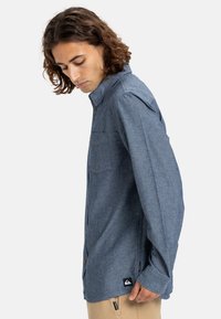 Button-up shirt in medium blue fabric, with long sleeves and a chest pocket. Features a contrasting black logo tag at the hem.