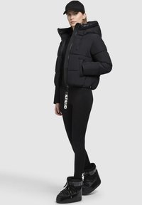 Svart pufferjacka med huva och framficka, bärs över svarta leggings. Tillbehören inkluderar en svart keps och glänsande svarta snöboots.