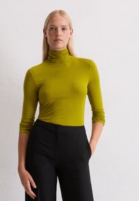 Langärmliger Rollkragenpullover aus leuchtend grünem Stoff, figurbetontes Design, mit einer glatten Textur und hohem Kragen, kombiniert mit schwarzen, maßgeschneiderten Hosen.