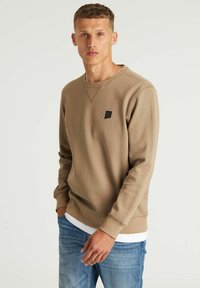 Sudadera beige con cuello redondo y mangas largas. Presenta un pequeño logo negro en el pecho y puños acanalados texturizados. Se lleva sobre una camisa blanca.