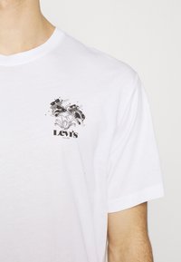 Valkoinen puuvillainen T-paita, jossa on musta graafinen painatus kukista ja kahdesta kalasta, alapuolella "Levi's" -teksti, lyhyet hihat ja pyöreä kaula-aukko.